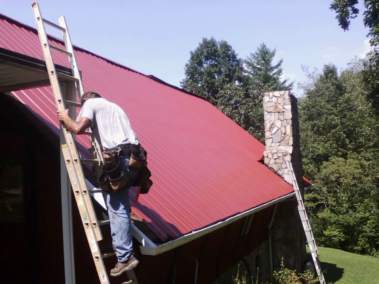 Roofer on a ladder beside a red metal roof and stone chimney — Soffit & Fascia Repair by Roofing Brothers in Skippack